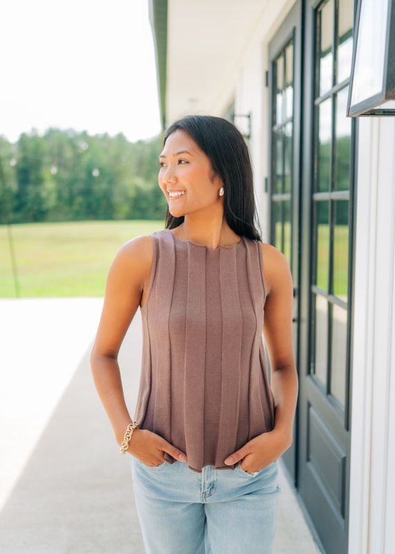 Side view of taupe sleeveless pleated knit tank styled with jeans, outdoor setting.