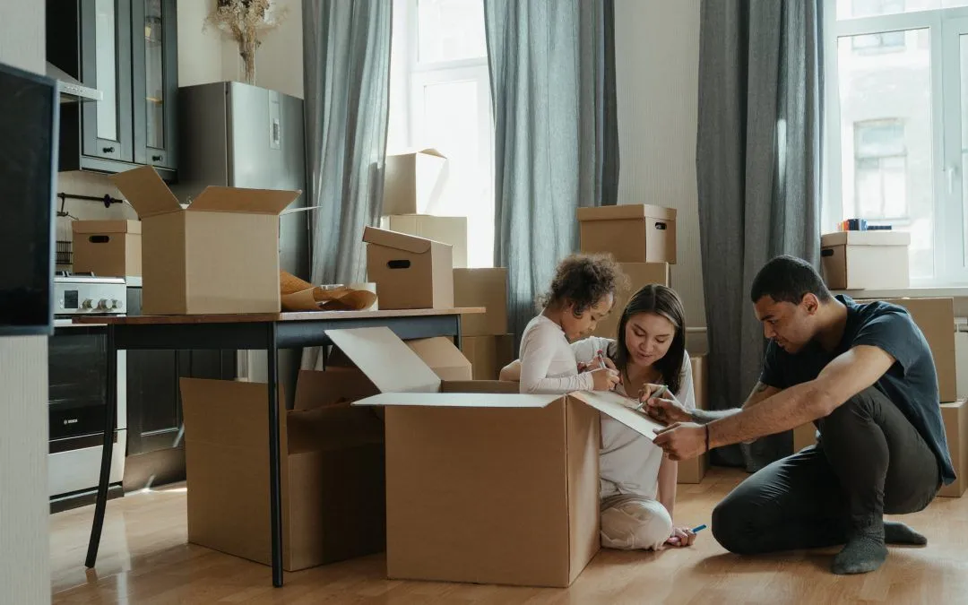 A family is sitting on the floor in a living room surrounded by cardboard boxes.