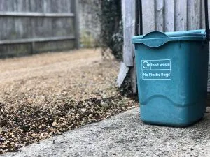 A blue trash can is sitting on the sidewalk next to a wooden fence.