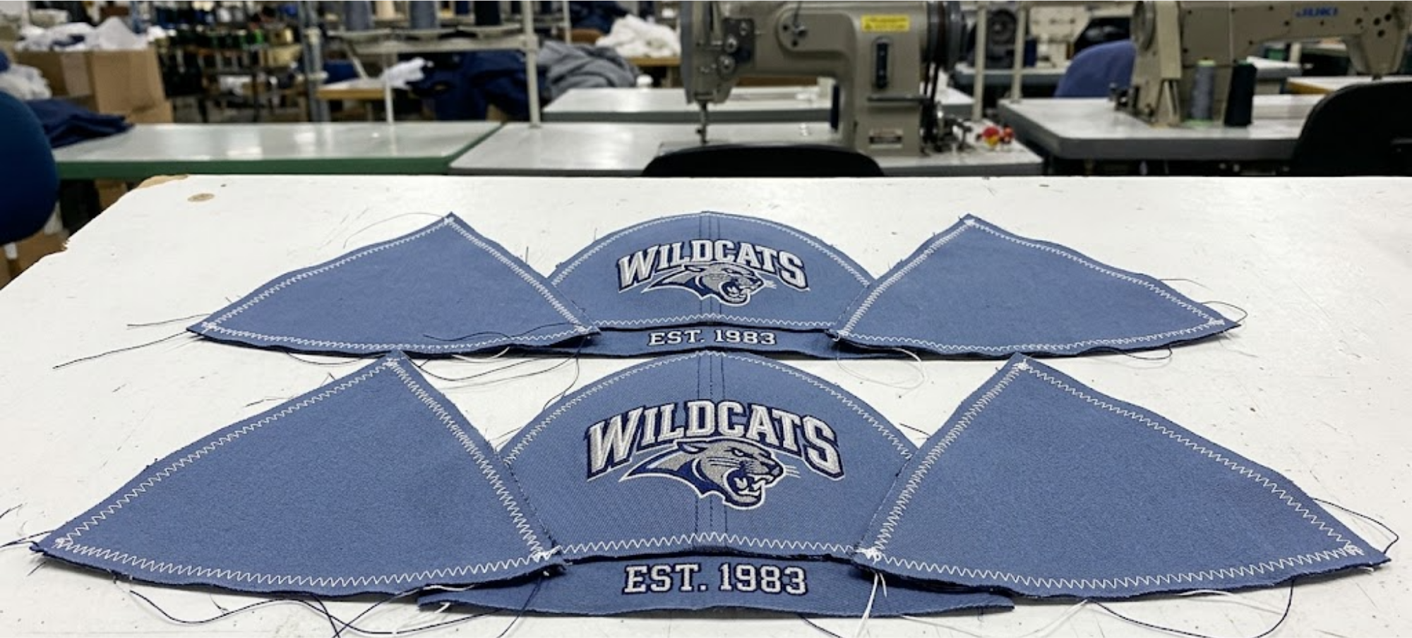 Unassembled baseball cap fabric panels laid out flat on a white background, showing embroidery across the seams and edges, industrial factory setting.