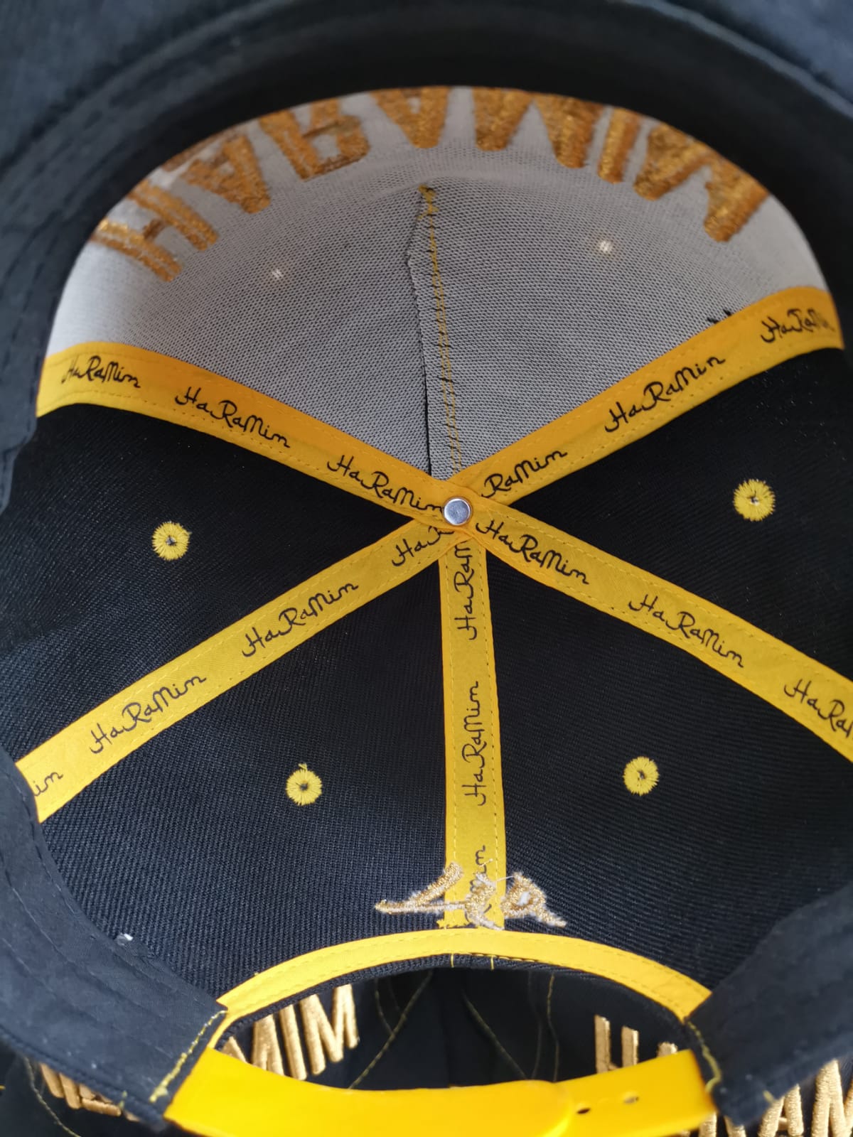 Underside view of a baseball cap showing black crown panels, yellow taping with branding along the seams, gold eyelets, and a yellow snapback closure.
